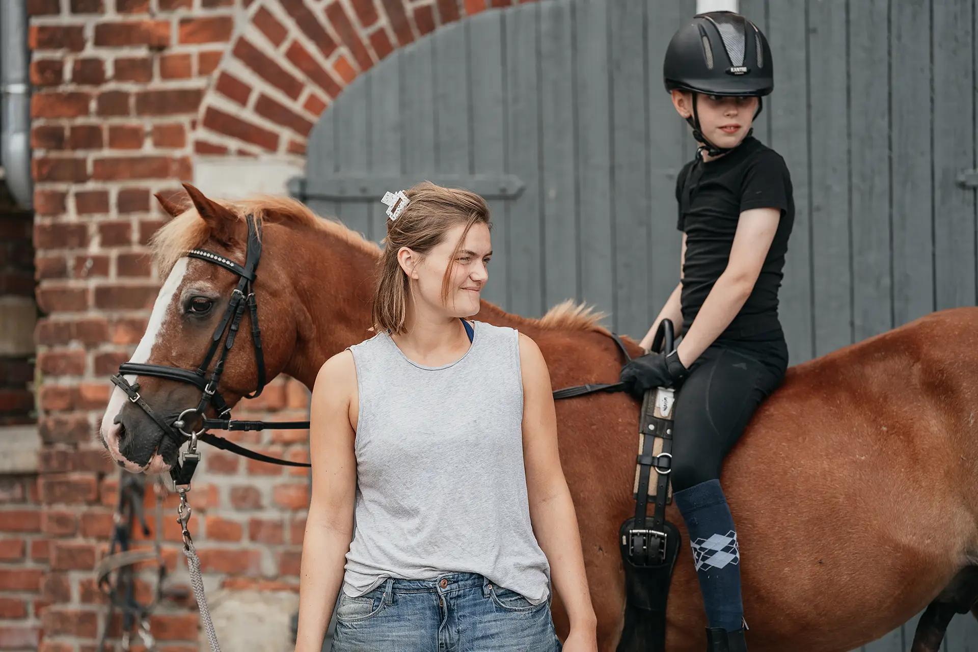 Ein Mädchen sitzt auf einem Pferd neben einer Betreuerin auf Ponyhof Mormann